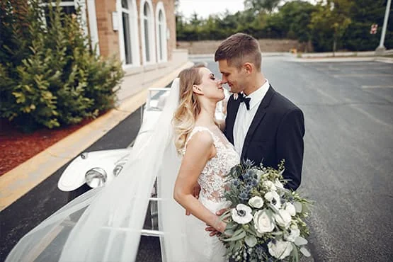 Un couple de mariés pose près d’une voiture blanche, avec un bouquet de fleurs.
