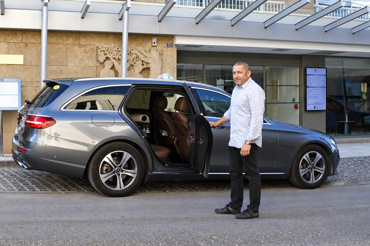 Un homme tient la portière arrière ouverte d’un taxi Mercedes gris devant un bâtiment.