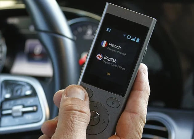 Une main tient un traducteur électronique affichant les langues française et anglaise dans une voiture.